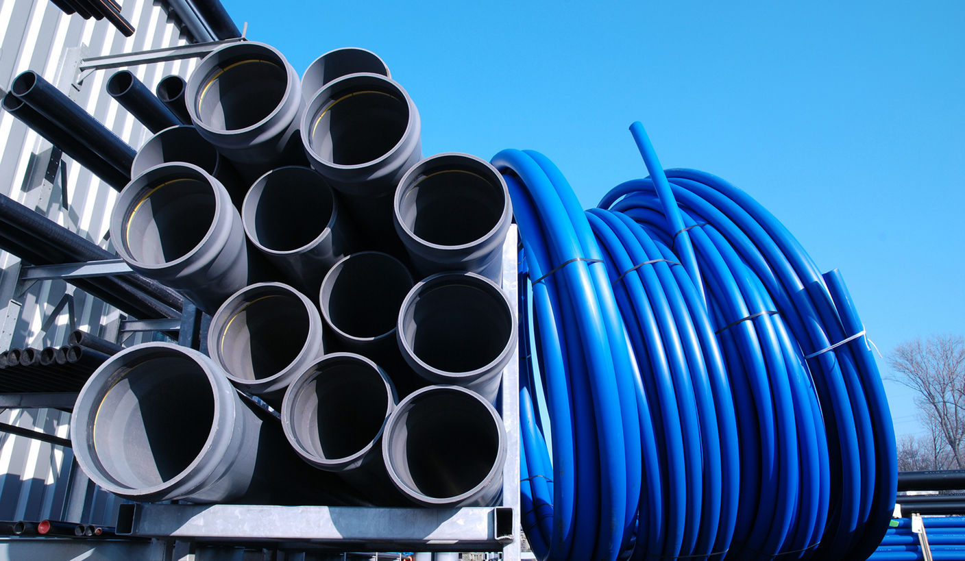An outdoor industrial site featuring a stack of large gray industrial pipes organized in a metal rack, alongside several large coils of blue tubing. The setting is under a clear blue sky, suggesting a construction or utility installation context.