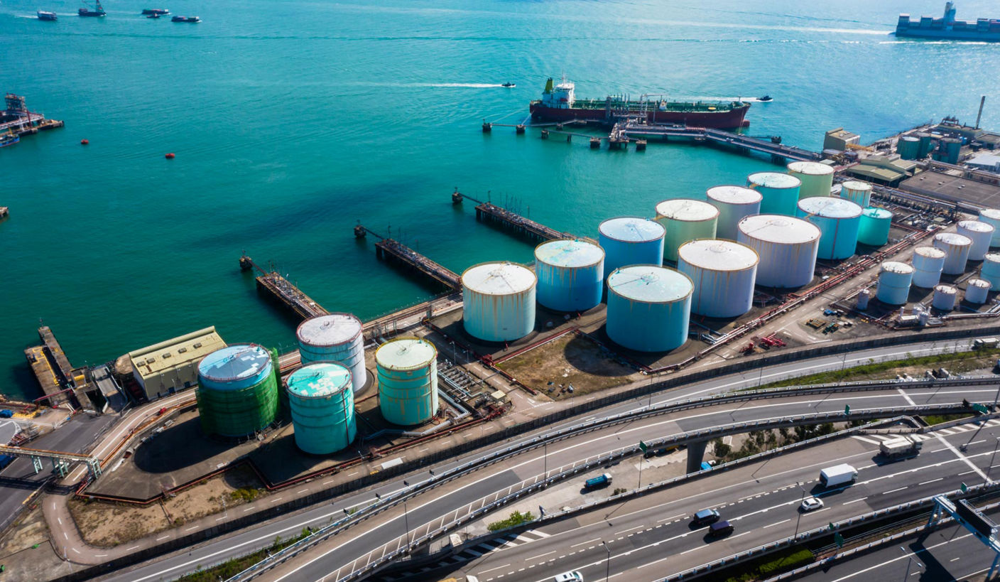 Aerial view of an industrial port with large cylindrical storage tanks, pipelines, and a tanker ship docked along a loading terminal in turquoise water.