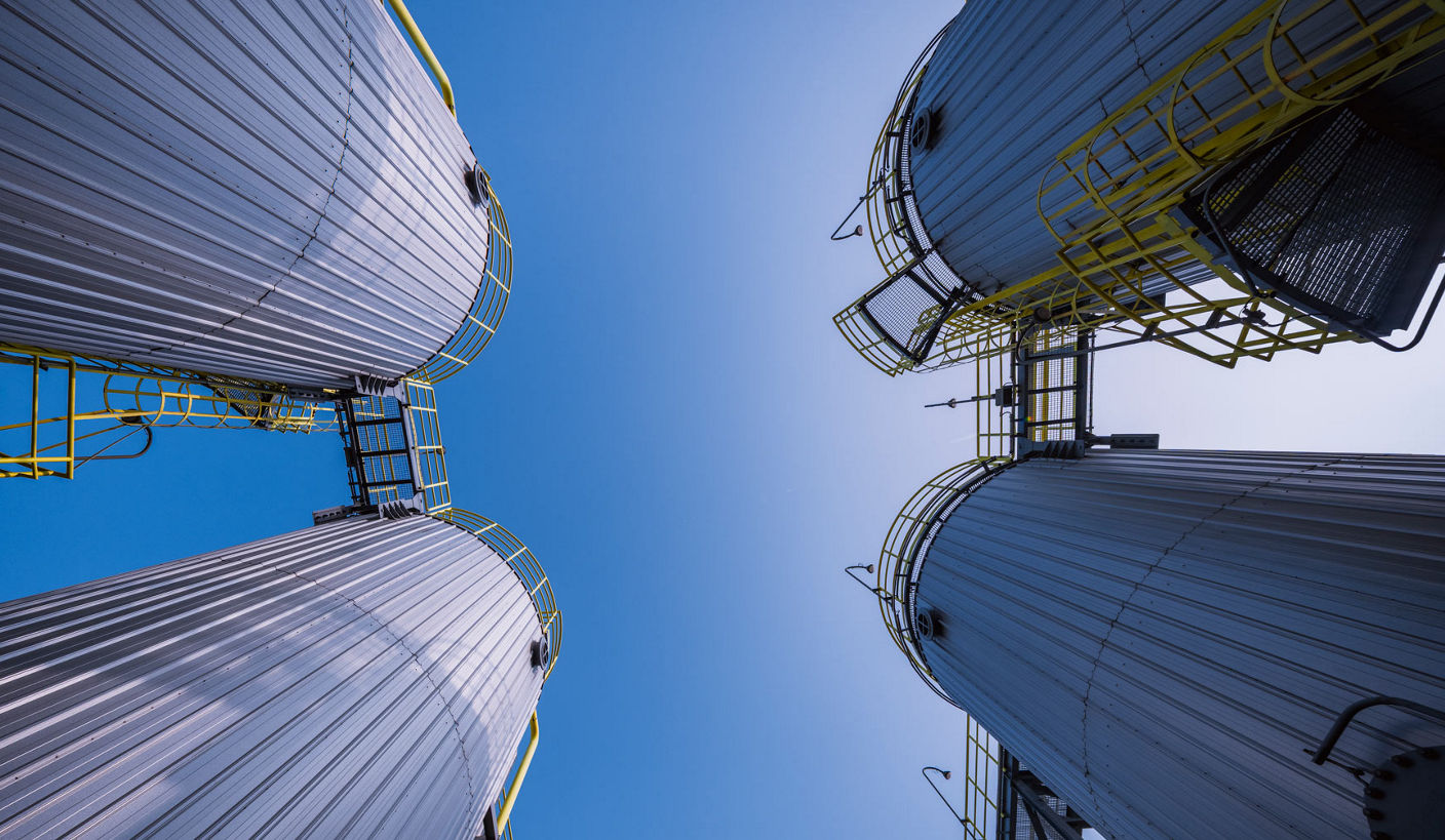 Upward view of tall industrial storage towers with ladders and platforms, framed against a bright blue sky.