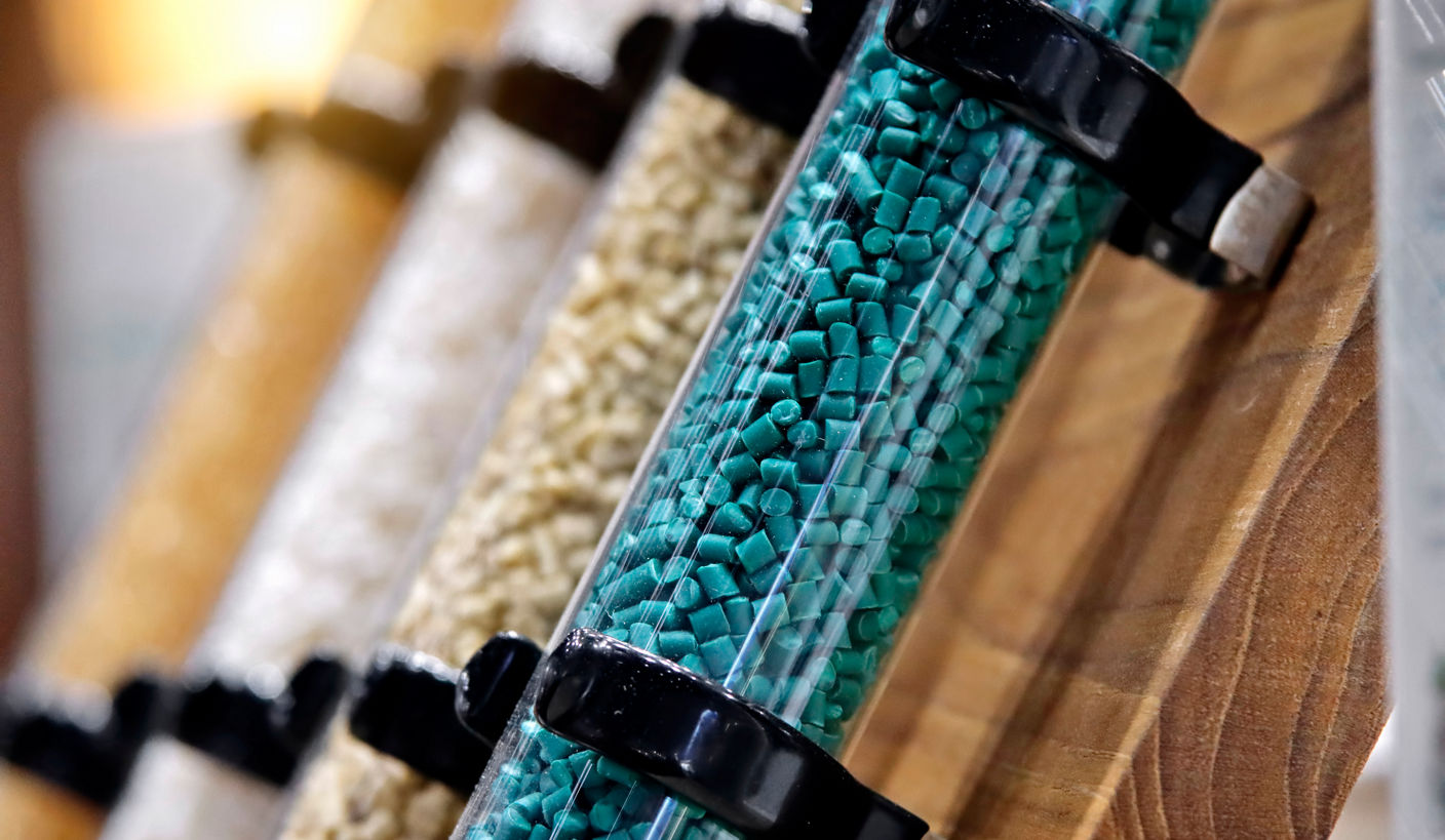 Row of transparent cylinders mounted on wood, each holding different colored plastic pellets, with teal granules in focus and beige and off‑white pellets behind, illustrating organized storage of raw manufacturing materials.