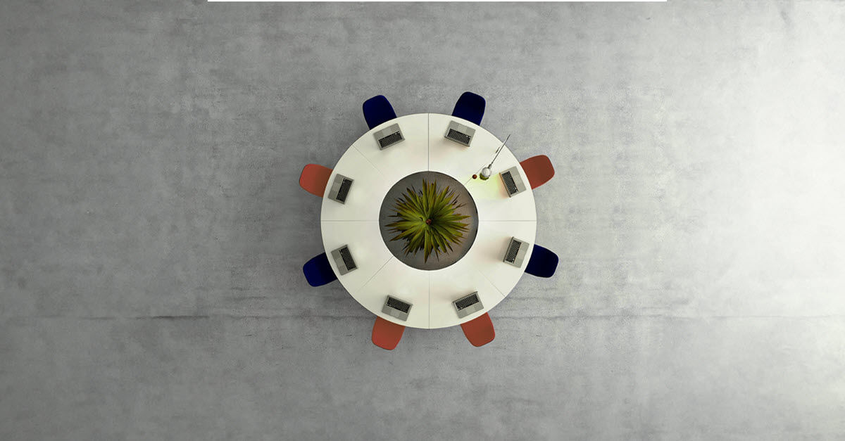 Top-down view of a round table with colorful chairs and laptops, centered by a plant.
