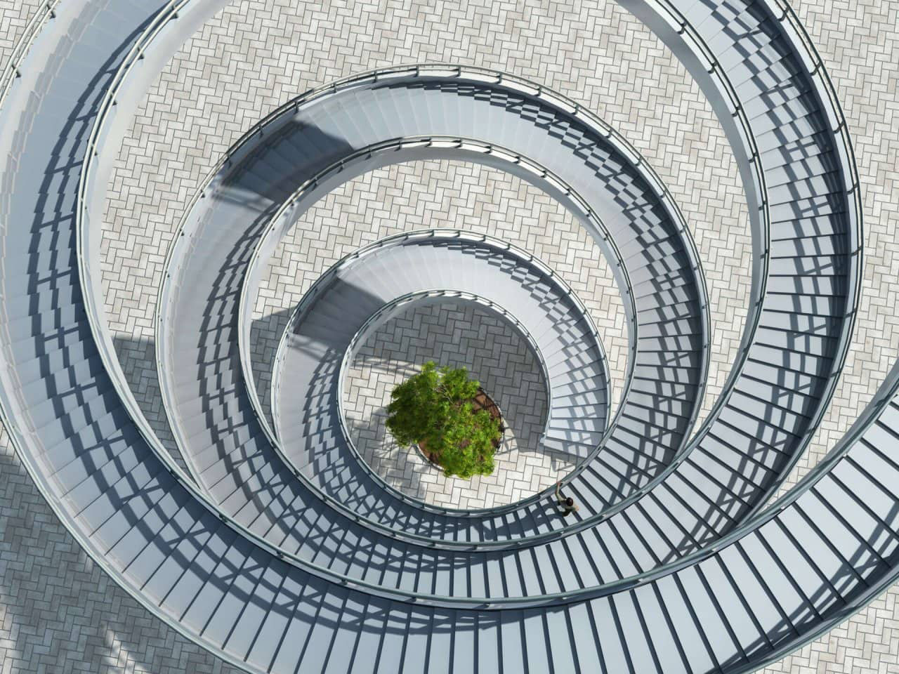 Aerial view of a spiral staircase with grey railings surrounding a small green tree at the center, set on a patterned pavement.
