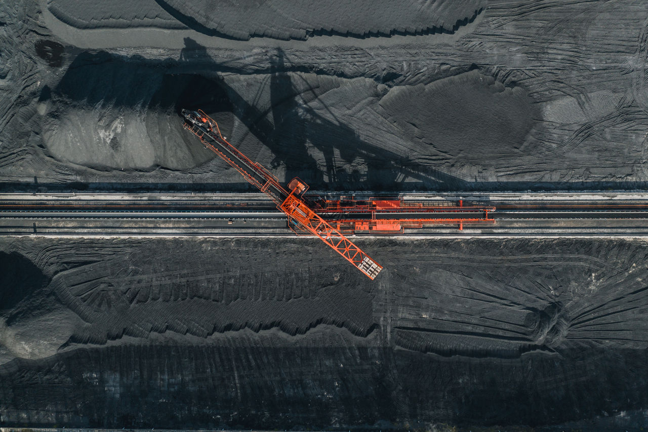 An aerial view of a large industrial machine, likely a conveyor or stacker, used in mining or material handling. The bright orange machine is positioned over tracks or conveyor belts, surrounded by dark material, possibly coal. Patterns indicate material movement, highlighting the scale and industrial nature of the operation.
