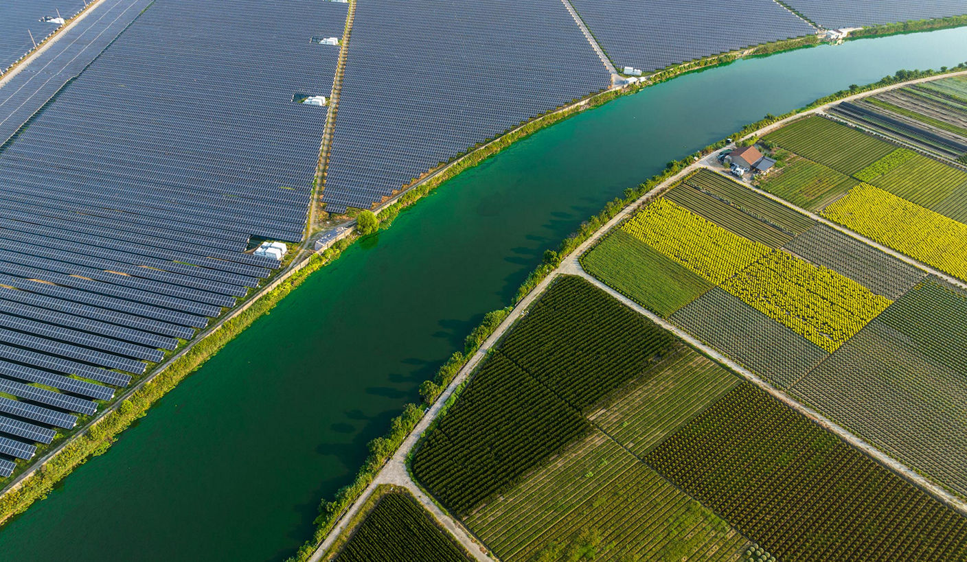 An aerial view shows a landscape divided into two main sections. On the left, a solar farm with solar panels arranged in a grid pattern occupies a significant area. On the right, diverse agricultural fields with various crops are visible, each with distinct colors and textures. A river or canal runs diagonally, separating the solar farm from the fields, with greenery along its banks. A small structure near the fields suggests a farmhouse or storage facility. The scene illustrates the coexistence of renewable energy infrastructure and agriculture.
