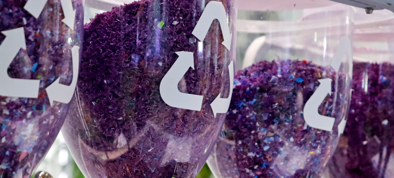 Transparent containers filled with small purple plastic pellets, each marked with a recycling symbol, represent processed recycled plastic
